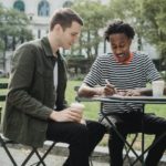 happy young diverse guys taking notes while doing assignment after studies in park